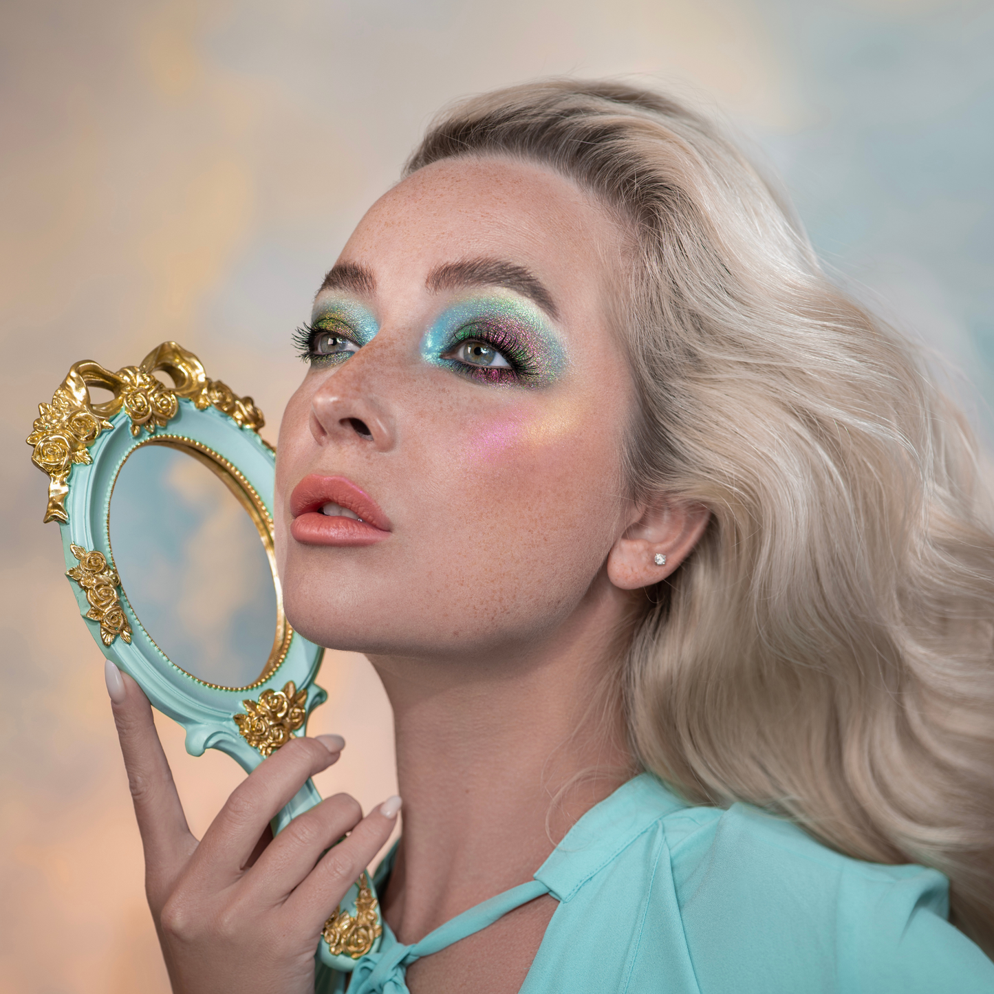 Woman with teal makeup holding a decorative mirror against a blurred background
